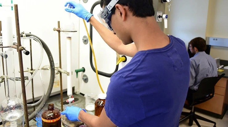 A student wearing safety goggles and gloves works with chemical physics equipment.