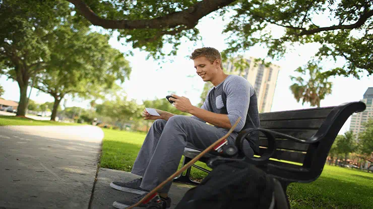 A student sits on a bench under a tree at Michigan State University, his longboard leaned on his backpack, as he smiles and looks at his phone